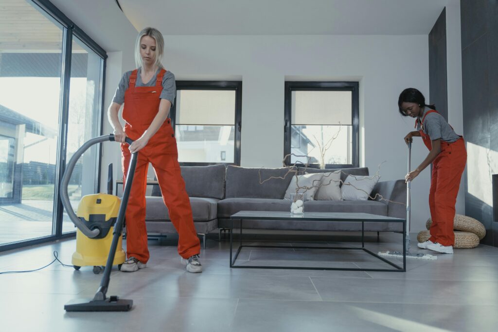 Home Two women in red overalls vacuuming and sweeping a modern living room.